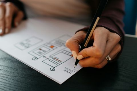 a woman's hand holding a pen, drawing a website wireframe on a piece of paper